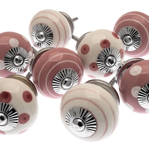Op de afbeelding: Een set van acht roze en witte keramische kastknoppen met zilveren accenten. De knoppen hebben verschillende patronen, waaronder stippen en strepen.