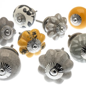 Könnte beinhalten: Sortiment an Keramik-Schrankknöpfen in Grau und Gelb mit silbernen Akzenten. Die Knöpfe haben verschiedene Muster und Texturen, darunter eine craquelée Glasur, florale Designs und Tiermuster.