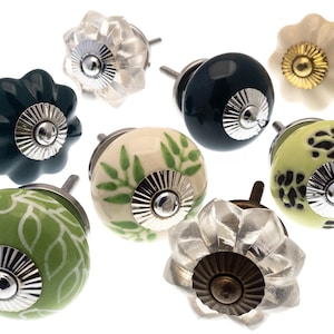 May include: Assortment of eight ceramic cabinet knobs in various colors and patterns. The knobs feature a green leaf design, a solid green color, a solid dark blue color, a cream color with green leaves, a light green color with black spots, and two clear glass knobs with a silver and gold finish.