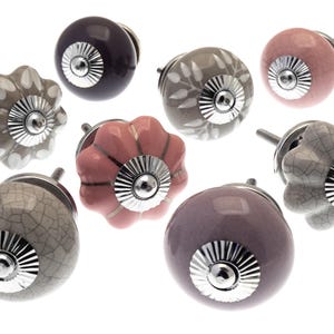 Könnte beinhalten: Sortiment an Keramik-Schrankknöpfen in verschiedenen Farben und Mustern, darunter rosa, grau und lila. Jeder Knopf hat einen silbernen Metallakzent.