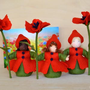 May include: Three felt dolls dressed in red jackets and green dresses, each holding a large red poppy flower. The dolls have different skin tones and hair colors. A colorful floral background is visible behind the dolls.