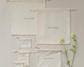 May include: A collection of six off-white fabric storage bags with drawstring or zipper closures. The bags feature gold text labels reading first outfit, first shoes, first teeth, hospital band, special things, and first haircut in a simple font.