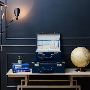 May include: Two navy blue vintage suitcases with gold accents and brass hardware. The top suitcase is open, revealing a white and blue polka dot lining. The suitcases are on a light wood table with a globe and a potted plant.
