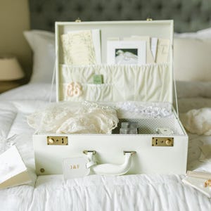 May include: A white vintage suitcase with gold accents and a luggage tag that says "J&F" is open and filled with white lace and paper goods. The suitcase is sitting on a white bedspread.