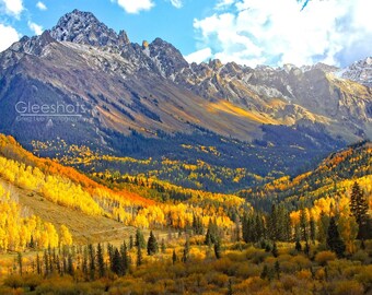 Puede incluir: Una cordillera con picos cubiertos de nieve y un valle lleno de follaje otoñal vibrante de color amarillo, naranja y rojo. El cielo es azul con nubes blancas.
