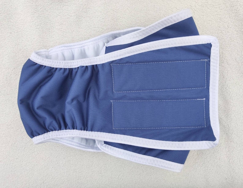 Op de afbeelding: Een herbruikbare hondenluier in blauw en wit met een witte rand. De luier heeft een klittenbandsluiting en is ontworpen om om de taille van de hond te worden gedragen.