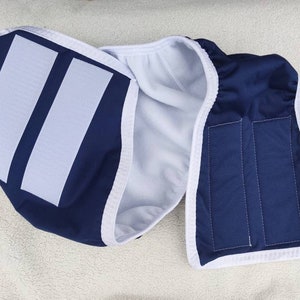 Op de afbeelding: Een herbruikbare blauwe en witte katoenen luier met twee witte klittenbandsluitingen. De luier heeft een witte fleece voering.