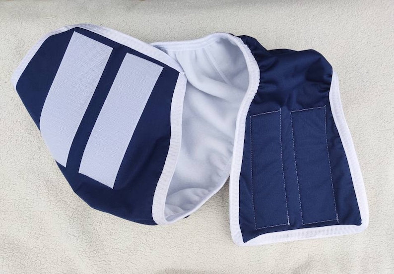 Op de afbeelding: Een herbruikbare blauwe en witte katoenen luier met twee witte klittenbandsluitingen. De luier heeft een witte fleece voering.