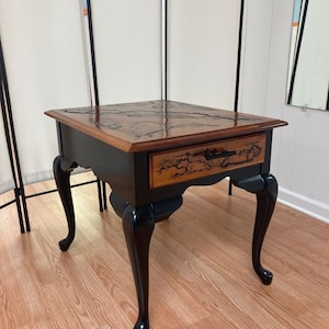 May include: A black end table with a wooden top featuring a unique wood grain pattern. The table has a single drawer and four cabriole legs.