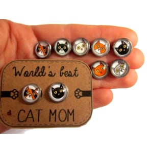 Puede incluir: Pendientes de botón con temática de gatos, con diseños de caras de gato variados, incluyendo gatos calicó, negros, blancos y naranjas. Los pendientes se muestran en una tarjeta marrón que dice "World's best CAT MOM".