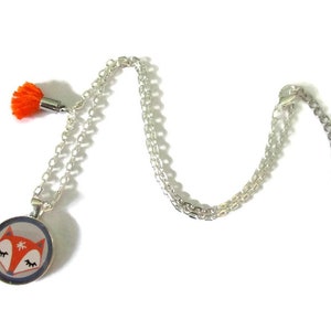 May include: Silver-toned necklace with a round pendant featuring a fox face design. The pendant has a white background, orange fox face, and a blue border. An orange tassel hangs from the chain. The necklace is on a white background.