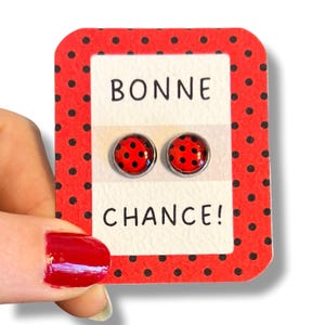 Könnte beinhalten: Ein Paar rote und schwarze Marienkäfer-Ohrstecker auf einer Karte mit dem Text "BONNE CHANCE!" in schwarz-weißen Buchstaben.