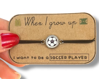 Könnte beinhalten: Eine braune Karte mit dem Text "Wenn ich groß bin, möchte ich Fußballspieler sein" und ein schwarzes Armband aus Schnur mit einem silbernen Anhänger in Form eines Fußballs.
