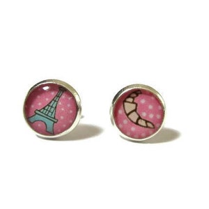Puede incluir: Un par de pendientes de plata con fondo de lunares rosa y blanco. Un pendiente tiene una Torre Eiffel azul y el otro un croissant marrón.