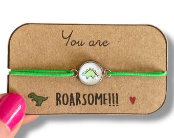 May include: Green elastic bracelet with a silver charm featuring a cartoon dinosaur. The bracelet is displayed on a brown card with the text "You are ROARSOME!!"