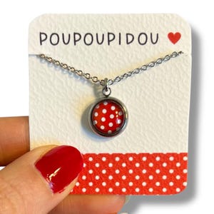 Könnte beinhalten: Eine silberne Kettenkette mit einem roten und weißen Tupfen-Anhänger. Der Anhänger ist rund und hat einen silbernen Rand. Die Kette wird auf einer weißen Karte mit dem Text "POUPOUIDOU" und einem roten Herzen präsentiert.