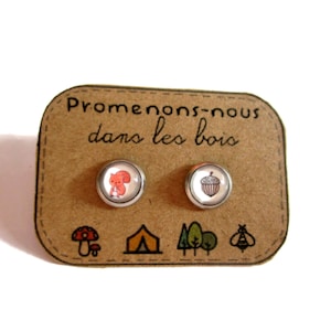 Könnte beinhalten: Runde Ohrstecker mit einem roten Eichhörnchen und einem Eicheldesign. Sie sind auf einer braunen Karte mit dem Text "Promenons-nous dans les bois" und Waldillustrationen abgebildet.