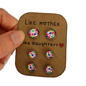 May include: Six colorful stud earrings on a brown card. The earrings have a mosaic design with various colors. The card reads "Like Mother Like Daughters" with a heart symbol. The earrings are held by a hand.