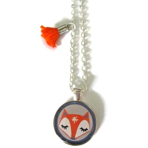 May include: A silver chain necklace with a round pendant featuring a cartoon fox face in orange, white, and gray. An orange tassel hangs from the chain. The pendant has a blue border.