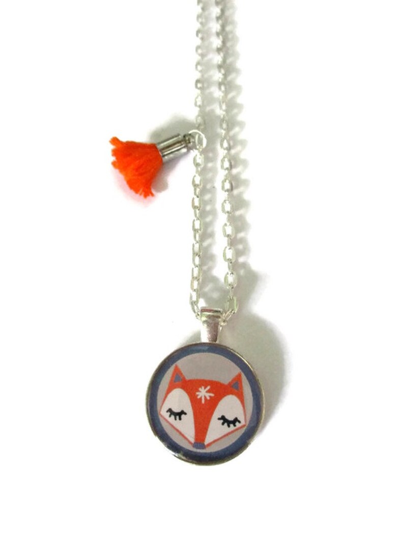 May include: A silver chain necklace with a round pendant featuring a cartoon fox face in orange, white, and gray. An orange tassel hangs from the chain. The pendant has a blue border.