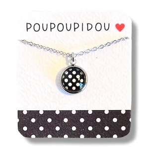 Puede incluir: Un collar de cadena de plata con un colgante de lunares blanco y negro. El colgante es redondo y tiene un borde plateado. El collar está sobre una tarjeta blanca con el texto "POUPOUIDOU" y un corazón rojo.