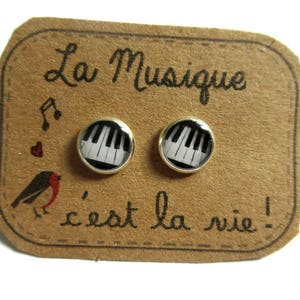 Peut inclure: Boucles d'oreilles à clous argentées avec un motif de clavier de piano. Elles sont présentées sur une carte marron avec le texte français "La Musique c'est la vie !" et une illustration d'oiseau.