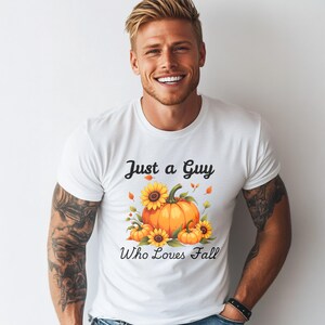 Pode incluir: Camiseta branca com o texto "Just a Guy Who Loves Fall" e um gráfico de abóboras e girassóis. O design apresenta abóboras laranja, girassóis amarelos e folhas de outono.