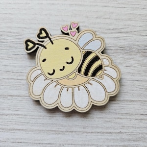 May include: A wooden cutout of a cartoon bee with a yellow body, black stripes, and a white flower. The bee has a happy face and is surrounded by pink hearts.