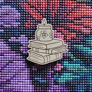 May include: A wooden cutout depicting a stack of books topped with a candle in a jar with floral designs. The background is a mosaic of small, round beads in purple, red, and blue hues.