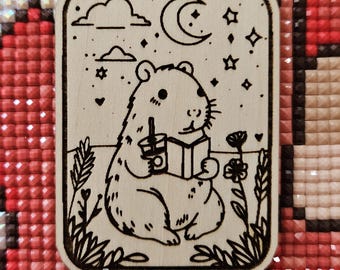 May include: A wood-burned design of a small animal holding a book and a cup, with a crescent moon and stars in the background.