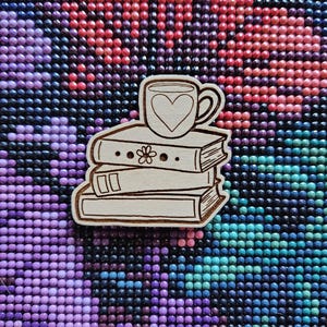 May include: A wooden pin depicting a stack of books topped with a coffee cup. The cup has a heart design. The books feature a flower and dot design. The pin is made of light brown wood.