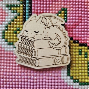 May include: A wooden cutout of a small dragon sleeping on a stack of books. The dragon has closed eyes and a small heart on the bottom book.