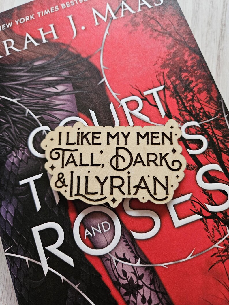 May include: A wooden bookmark with the text "I like my men's tall, dark & Illyrian" on a red and black book cover with the text "A Court of Thorns and Roses".
