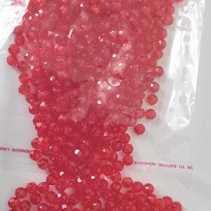 May include: A clear plastic bag filled with numerous faceted, red beads. The beads are small and have a shiny, reflective surface. The bag has text that reads "REMOVE LINE" and "ER TO EXPOSE ADHESIVE".