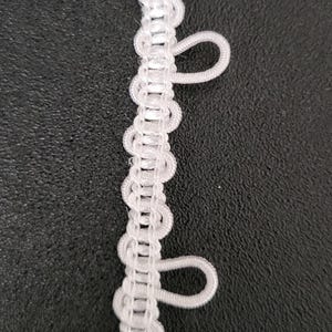 May include: Close-up of white loop trim. The trim features a series of small loops along one edge, connected by a woven center. The loops are evenly spaced and create a decorative edge. The trim is against a dark background.