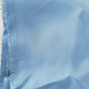 May include: A close-up of a light blue fabric, possibly cotton or linen, with a raw, unfinished edge. The fabric is folded, showing the texture and color variations. The color is a soft, pastel blue.