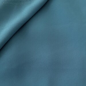 May include: A close-up shot of a teal fabric with a subtle texture. The fabric is folded over, revealing a perforated edge. The color is a muted, cool tone, and the material appears to be soft and pliable. The image highlights the fabric's smooth surface and slight sheen.