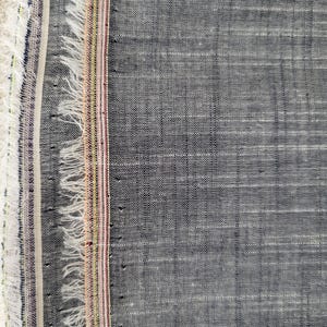 May include: Close-up of a textured, gray fabric with a frayed edge. The fabric has a woven appearance with subtle horizontal and vertical lines. A colorful striped border with white, purple, and yellow accents runs along the left edge.