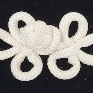 May include: Ivory-colored frog closure made of woven cord. The closure features a central knot with looped ends, creating a decorative and functional element. Ideal for garments, this closure adds a touch of elegance.