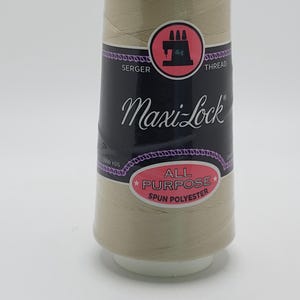 May include: A spool of beige Maxi-Lock all-purpose spun polyester serger thread. The black label features a pink circle with a black serger machine icon, the text Maxi-Lock, and the words All Purpose Spun Polyester with 3000 YDS printed on it.
