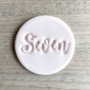 Könnte beinhalten: Ein transparenter Stempel aus Kunststoff mit dem Wort "Seven" in einer kursivschriftlichen Schriftart. Der Stempel befindet sich neben einer rosa Tonscheibe mit dem eingeprägten Wort "Seven".