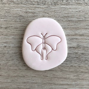 Può includere: Un timbro in argilla polimerica rosa con un design a forma di farfalla. La farfalla è delineata in una tonalità di rosa più scura.
