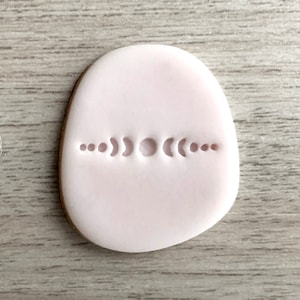 Può includere: Uno strumento in plastica bianca con una fila di fasi lunari, utilizzato per creare impressioni in fondant o argilla. Lo strumento è accanto a un biscotto di fondant rosa con un design di fasi lunari.