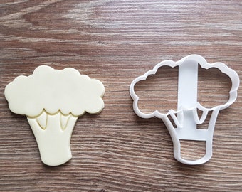 May include: A white plastic cookie cutter shaped like a broccoli floret. The cutter is shown next to a yellow cookie that has been cut out using the cutter.