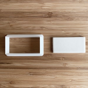 Puede incluir: Dos cortadores de galletas de plástico blanco, uno con forma de rectángulo y el otro con forma de cuadrado.