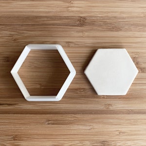 Puede incluir: Dos cortadores de galletas hexagonales de plástico blanco, uno con una forma sólida y el otro con un contorno. Los cortadores están sobre una superficie de madera.