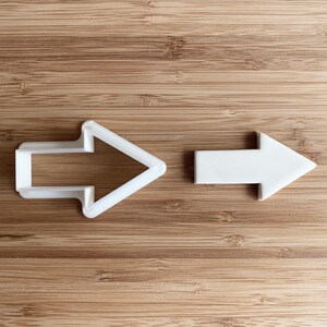 Puede incluir: Dos cortadores de galletas en forma de flecha de plástico blanco sobre una superficie de madera. La flecha de la izquierda es más detallada que la flecha de la derecha.
