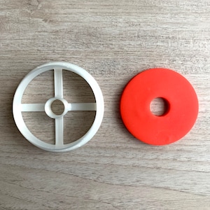 Puede incluir: Un cortador de galletas de plástico blanco en forma de círculo con cuatro radios y un círculo más pequeño en el centro. Junto a él hay un cortador de galletas en forma de rosquilla roja con un agujero en el centro.