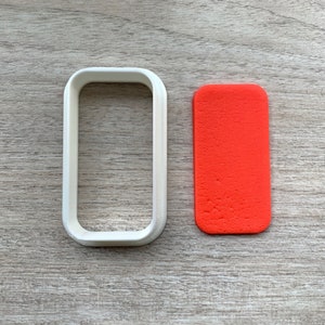 Puede incluir: Un cortador de galletas de plástico blanco en forma de rectángulo, junto a un rectángulo rojo de fondant.