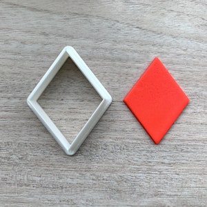 Op de afbeelding: Een witte plastic diamantvormige koekjesvormer en een rode diamantvormige koek.